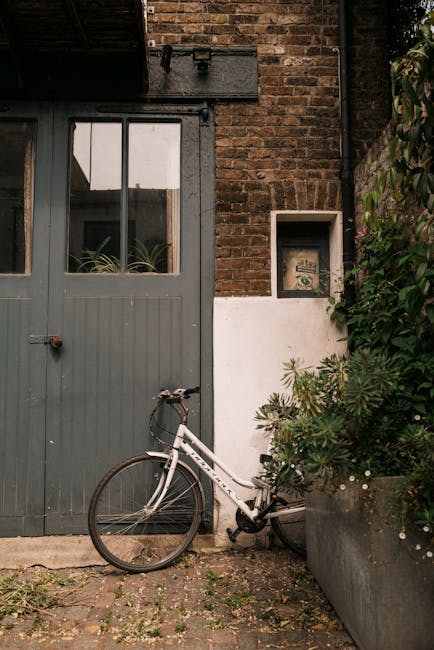 A close-up view of an urban garden corner featuring a large, weathered gray wooden door with a small, rectangular window at the top, framed by a brick wall. Beside the door, a white bicycle with black