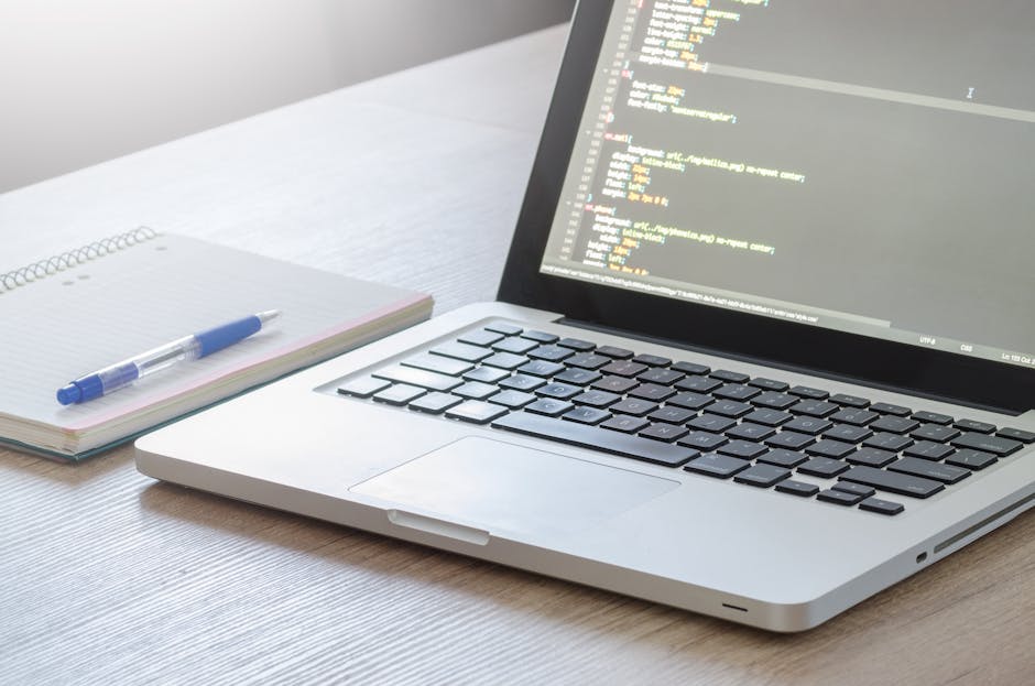A close-up of a modern silver laptop with a black keyboard displaying lines of colorful code on its screen, situated on a light wooden desk. To the left of the laptop, there is a spiral-bound notebook