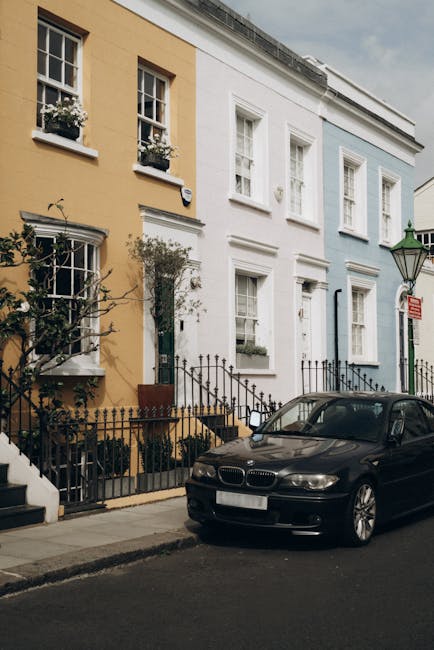 A row of colorful terraced houses on Portobello Road, featuring facades painted in yellow, white, and light blue, with tidy window boxes filled with white flowers on the upper floors. The ground-level