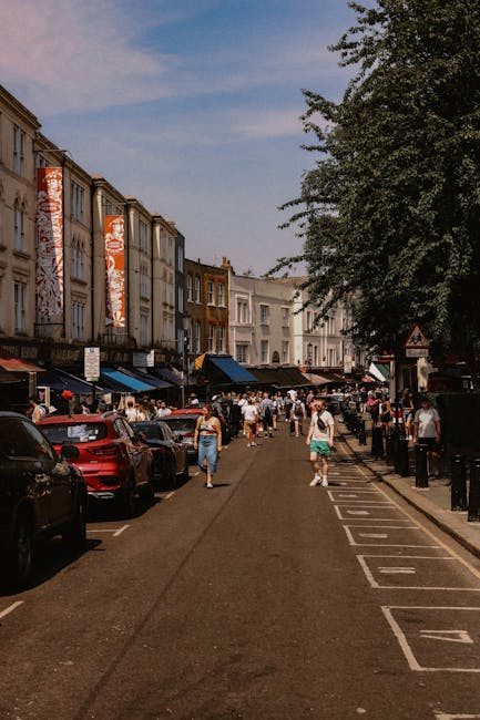 A row of colorful terraced houses on Portobello Road, featuring facades painted in yellow, white, and light blue, with tidy window boxes filled with white flowers on the upper floors. The ground-level